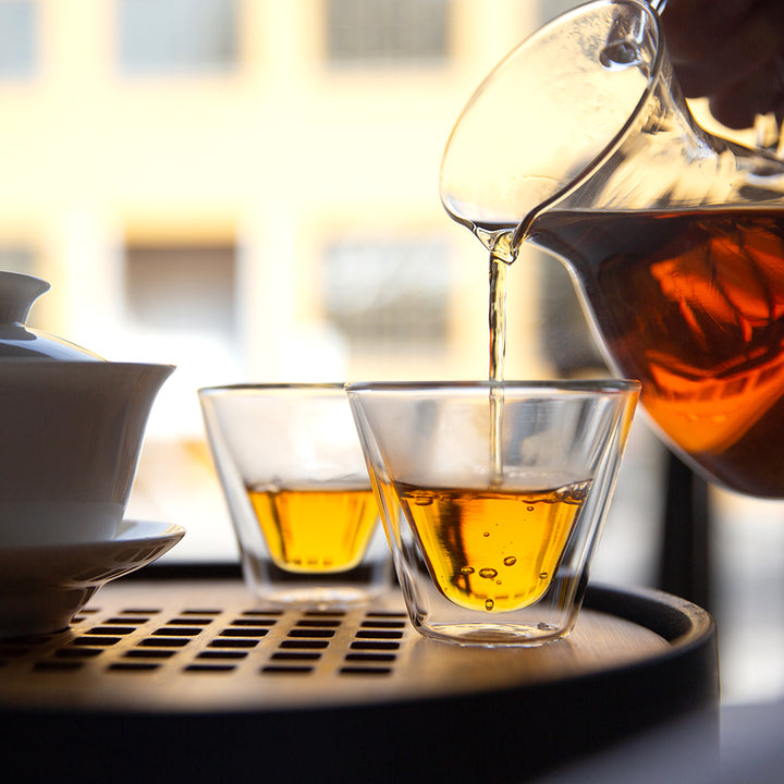 Glass tea decanter pouring tea into two small glass gaiwan cups.