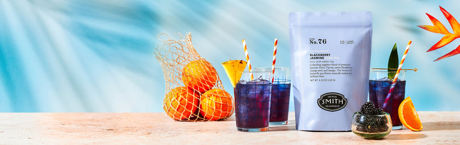 Bag of Blackberry Jasmine iced tea beside three glasses of brewed blue iced tea and a bag of oranges.