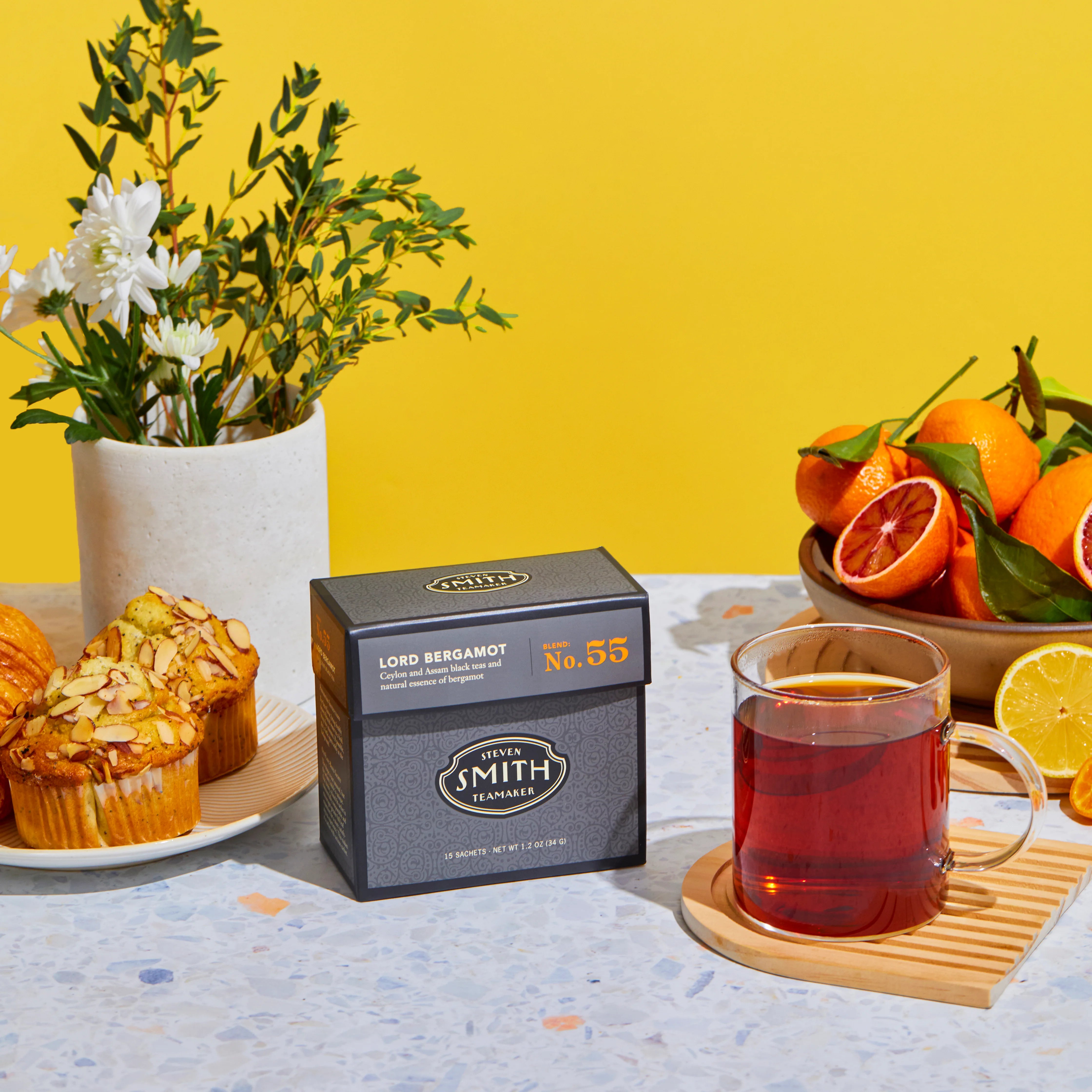 Grey box of Lord Bergamot black tea on a table next to a brewed cup of tea, a bowl of citrus and a plate of muffins.