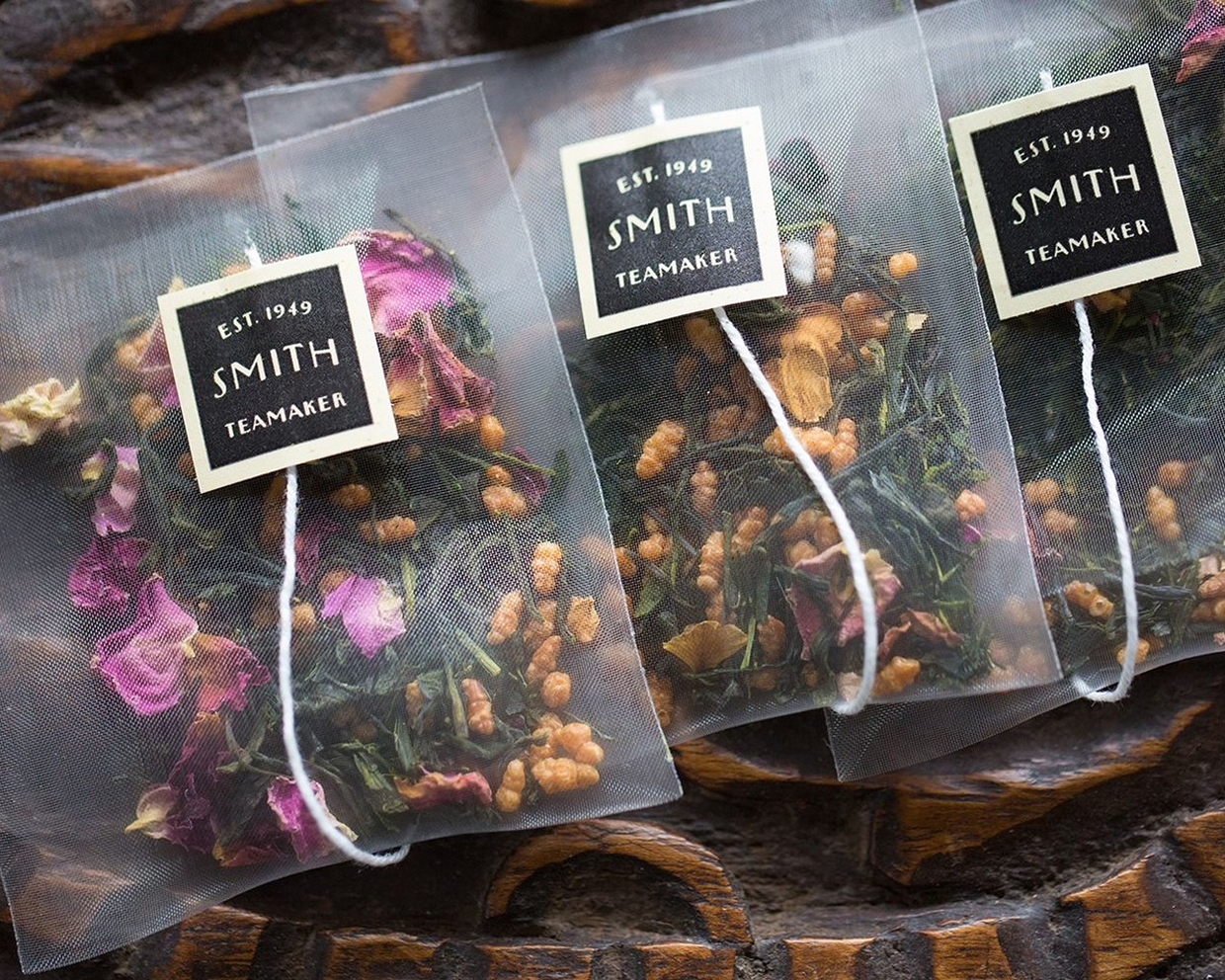 Three clear tea bags with white strings and cream labels reading 'SMITH TEAMAKER EST. 1949' are arranged on a rustic dark wood surface.