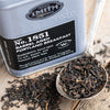 Silver spoon on a wooden table filled with loose black tea next to silver tin with a black label.