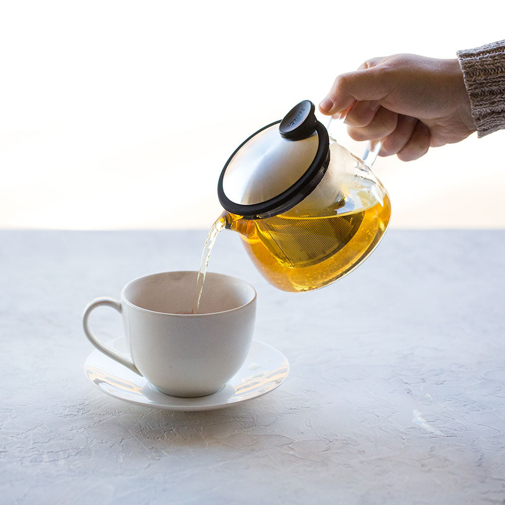 Glass teapot with stainless steel and black lid with spout and handle filled with amber colored tea.