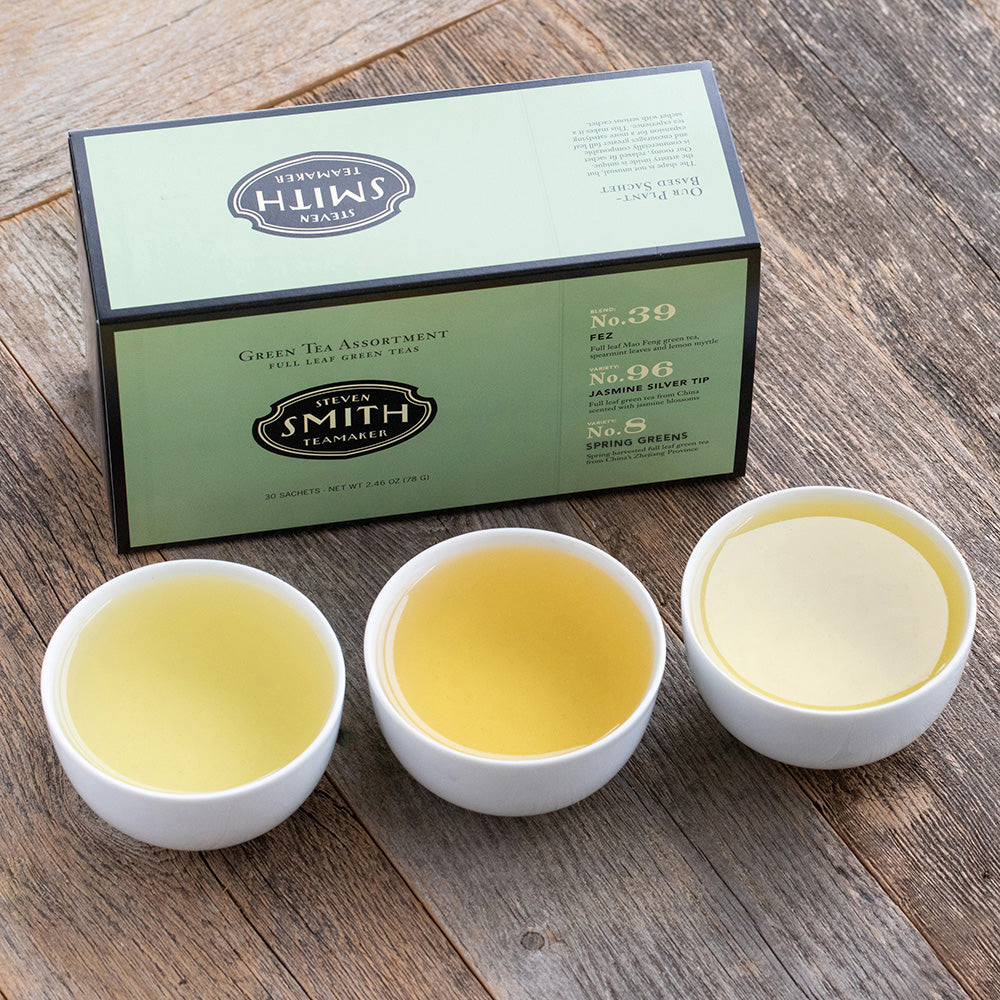Green box with black trim on wooden table next to three porcelain cups filled with green teas.